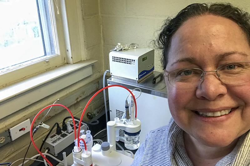 Genevieve Bernatchez wears glasses and a striped shirt in a selfie of her in the lab. There is analytical chemistry equipment in the background