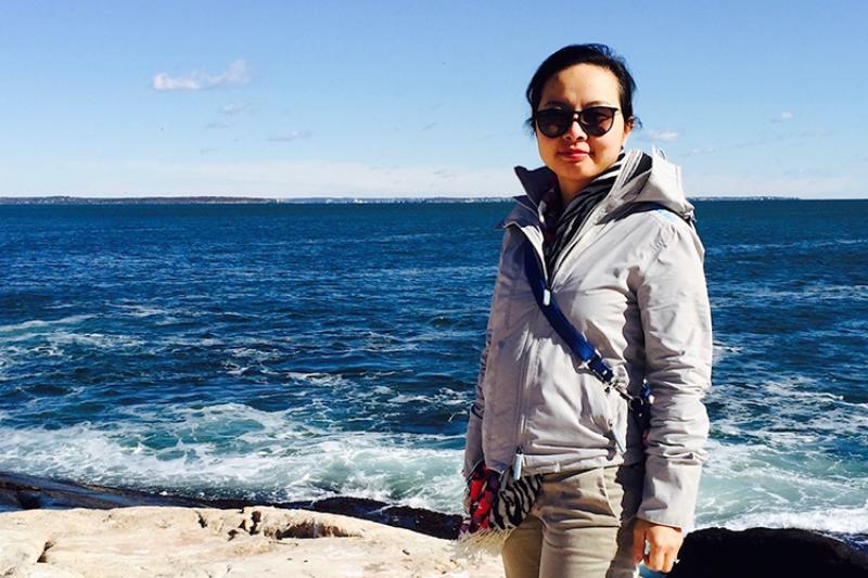 Changhua Weng wears sunglasses and a gray jacket while standing on a rocky coastline.