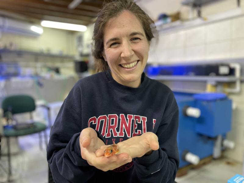 A woman in a dark blue sweater holds a small orange fish in her open palms.