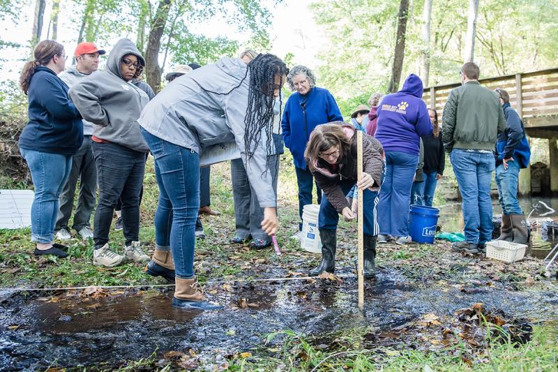 Two women lean over to examine how one of the women is holding a yardstick in a stream as other people look on. Credit: Virginia Commonwealth University.