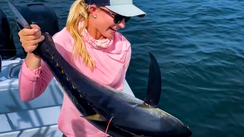 A woman in a hat and glasses on the deck of a boat holds a tuna with a red, string-like object attached near its top fin
