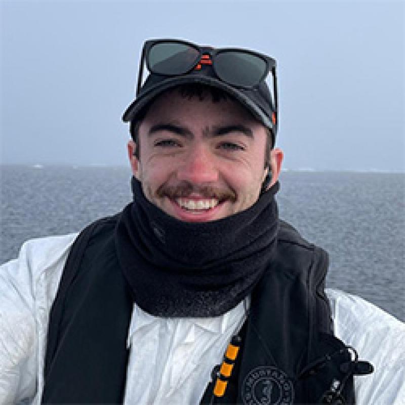 Portrait of scientist with sunglasses and hat in black scarf and wearing a white shirt, background is the ocean