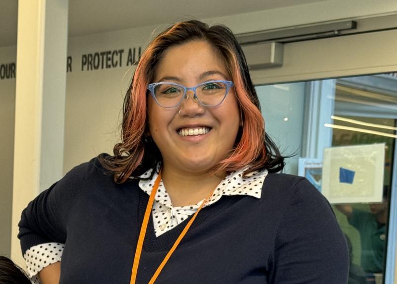A woman in blue-rimmed glasses, a black sweater and white shirt with black polka dots smiles at the camera.