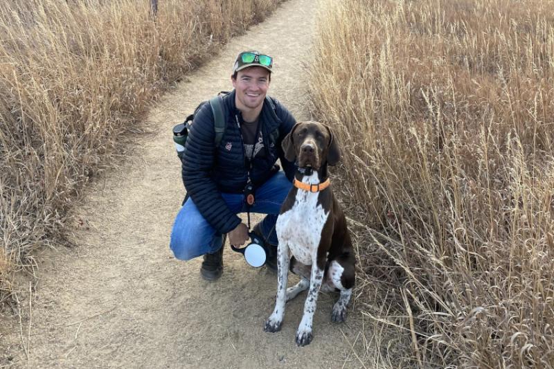 A young man squats next to a large dog on a trail.