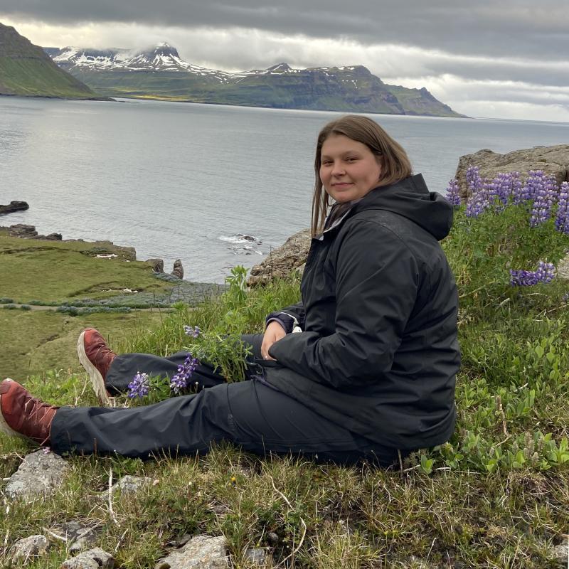 Abigail Lucas sitting above ocean, purple lupine behind her. 