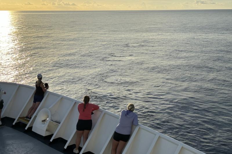 Three women stand at the bow of a ship looking a several circular, calm patches of water in a row.