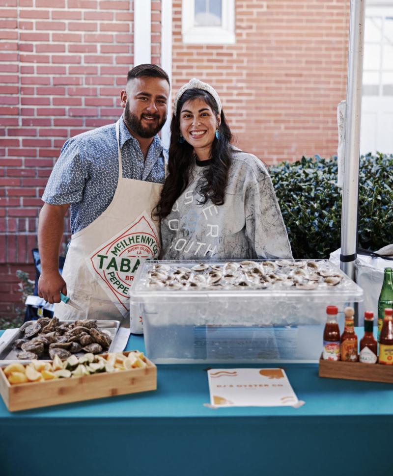 A married couple stand arm in arm, smiling, in front of an oyster booth with shucked oysters and garnishes. 
