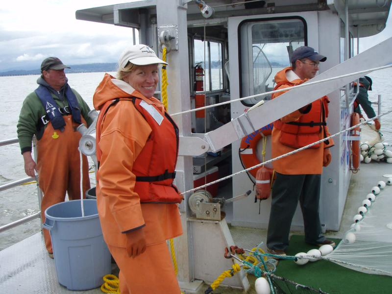 Three people standing on deck of a ship, with woman as central figure. She's wearing orange jacket, pants, gloves and a safety vest, and is half turned and smiling at the camera.