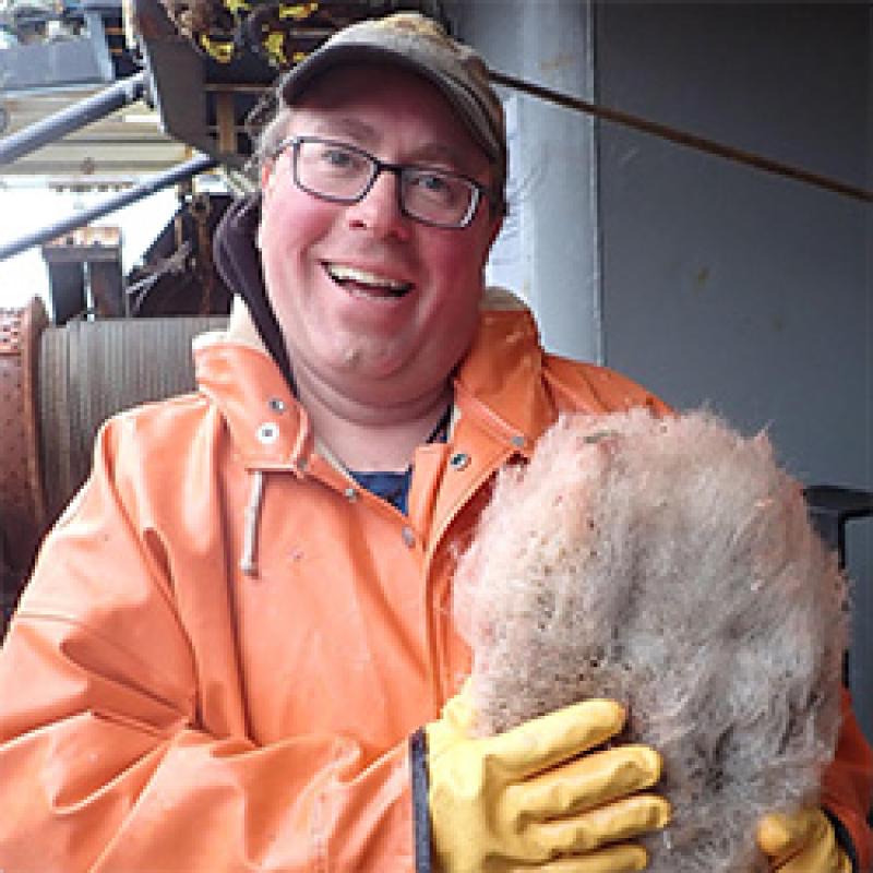 Scientist wearing orange jumpsuit, hat and glasses holding a sponge.