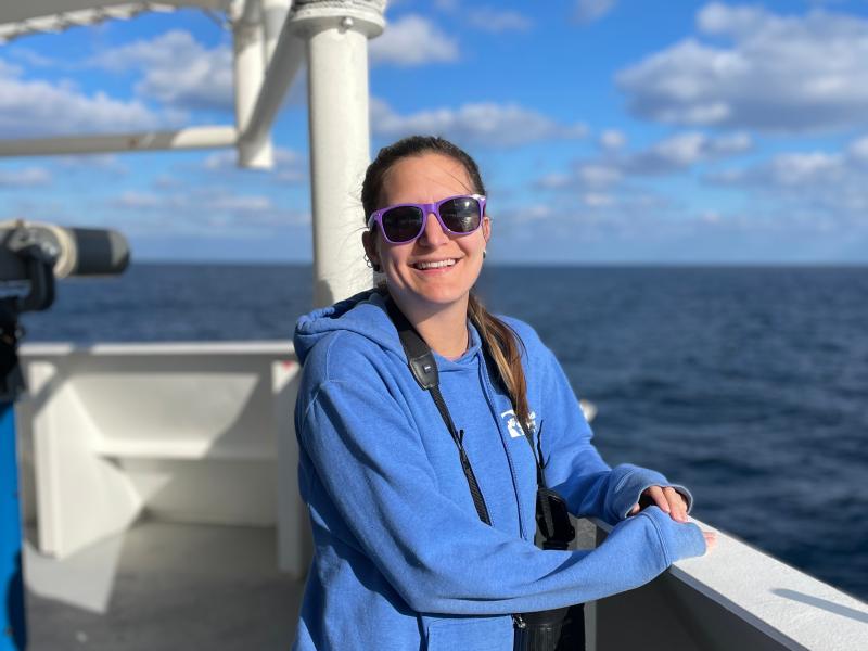 Kiersten stands at the taffrail of the ship and smiles for the camera. She is wearing a sweatshirt, purple sunglasses, and a pair of binoculars around her neck. the sky is blue with many clouds over a darker blue ocean.