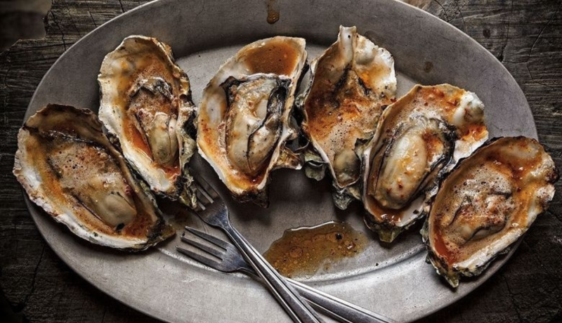 six cooked oysters on the half shell on a silver plate with two three-pronged forks.