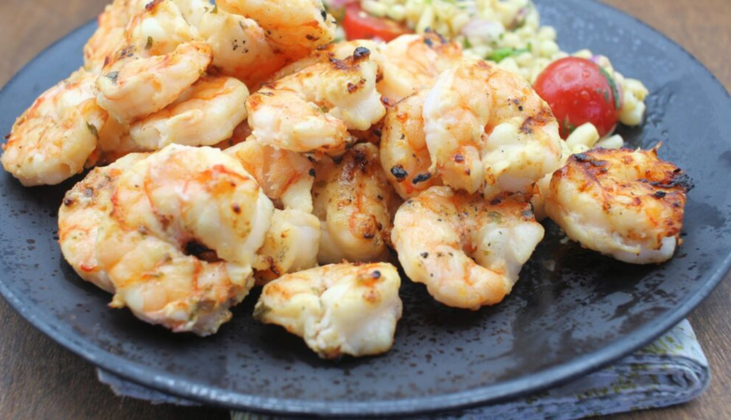 A side-view of a black plate with a mound of grilled shrimp on it. Tomatoes and rice are on the plate in the background.