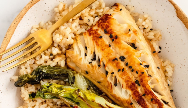 a birds-eye view of a white bowl speckled with black dots. in the bowl is rice, cooked vegetables, and a seared fish filet sprinkled with black sesame seeds. also in the bowl is a four-pronged gold fork.