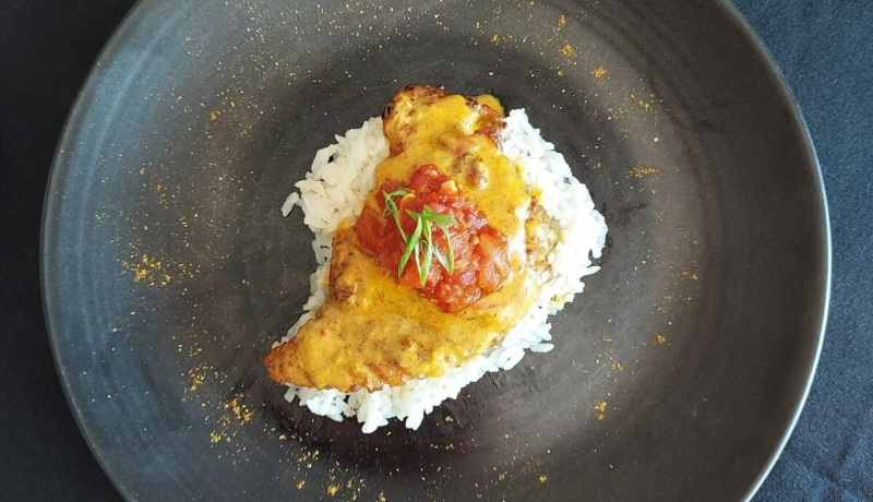 a birds-eye view of a black bowl. In the bowl is white rice with a cooked skate filet on top. the skate filet is topped with a yellow-orange curry sauce, red chutney, and green herbs.