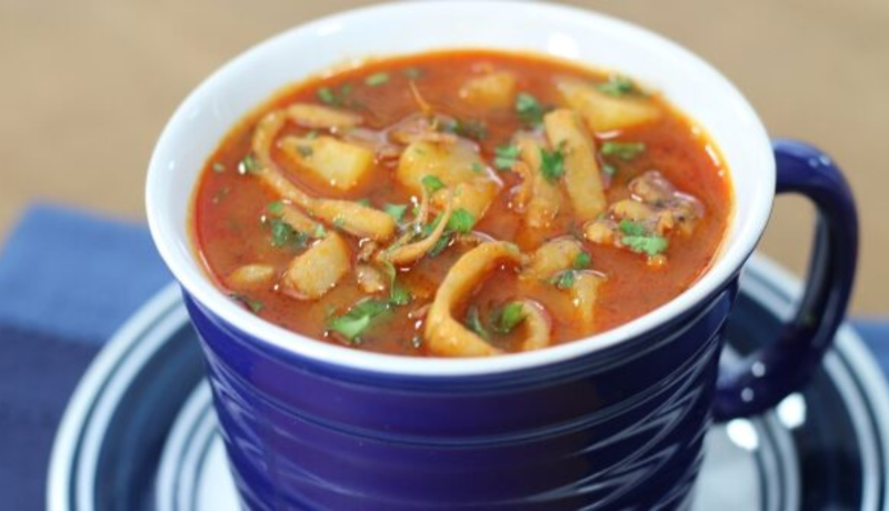 a large ceramic mug sitting on a tray. the outside of the mug is blue and the inside is white. it is filled with a red tomato broth squid chowder that is garnished with green herbs.