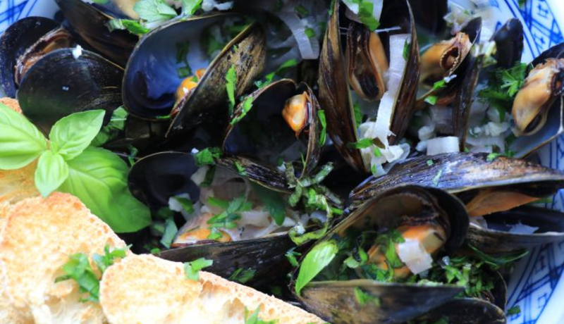 a birds-eye view of a bowl of cooked mussels with their shells open. the mussels have green leafy herbs sprinkled over them and some toasted oval-shaped bread slices.