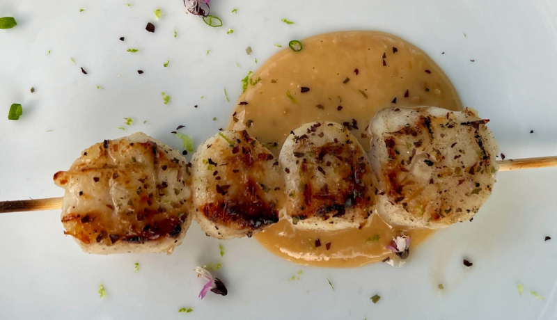 four seared scallops on a wooden skewer plated with a yeallow sauce and green and purple herbs.