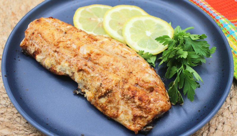 A dark blue plate with a grilled fish filet on it. The filet has a brown-red coating. Next to the filet are three round slices of lemon and some leafy green herbs.