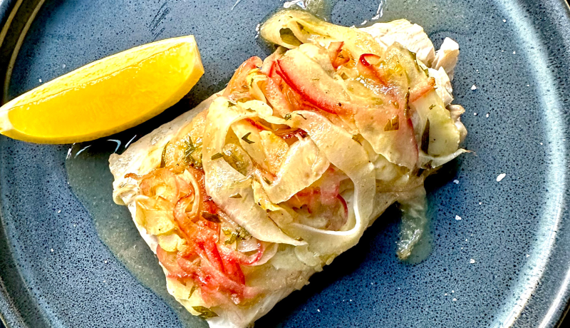 A blue and black speckled ceramic plate with a baked fish filet. The filet is topped with red, orange, and white cooked vegetables and seasoned with green herbs. Also on the plate is a lemon wedge.