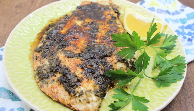 A light green and white ceramic plate sitting on a blue and white cotton napkin. On the plate is a cooked filet of flounder with a dark herb sauce on top. The plate is garnished with a lemon wedge and green leafy herbs.