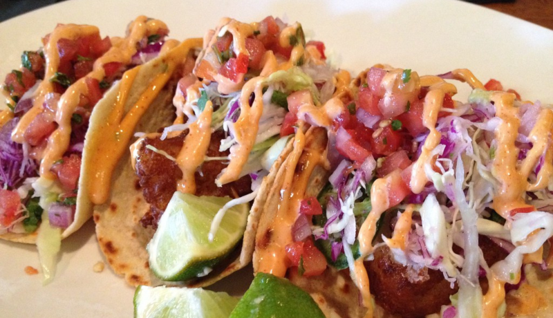 A plate of three tacos made with flour tortillas loaded with fried fish, chopped vegetables, and an orange aioli sauce. There are three small lime wedges on the plate.