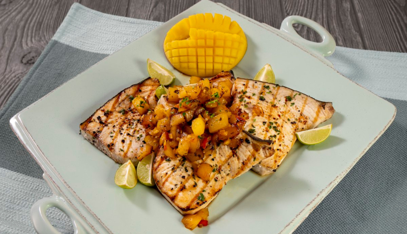 A light green square plate with small handles on two sides sitting on a wooden table with a cloth underneath it. On the plate are two grilled swordfish steaks topped with mango chutney. Surrounding the swordfish is a halved mango with the fruit cubed, and five lime wedges.