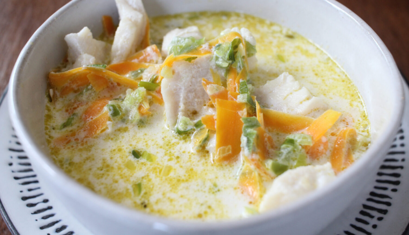 A white ceramic bowl on a blue and white plate. The bowl is filled with a creamy soup that includes white fish, greens, and cheese.
