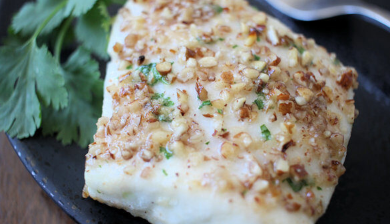 A black plate with a pecan-encrusted cooked white fish filet. The filet is also seasoned with green herb.
