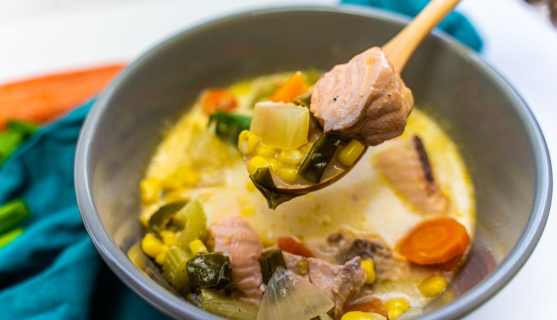 A grey ceramic bowl filled with a creamy chowder that has carrots, onions, celery, corn, cooked salmon chunks, and chopped herbs mixed in. There is a wooden spoon hovering above the bowl with a bite of the chowder in it.