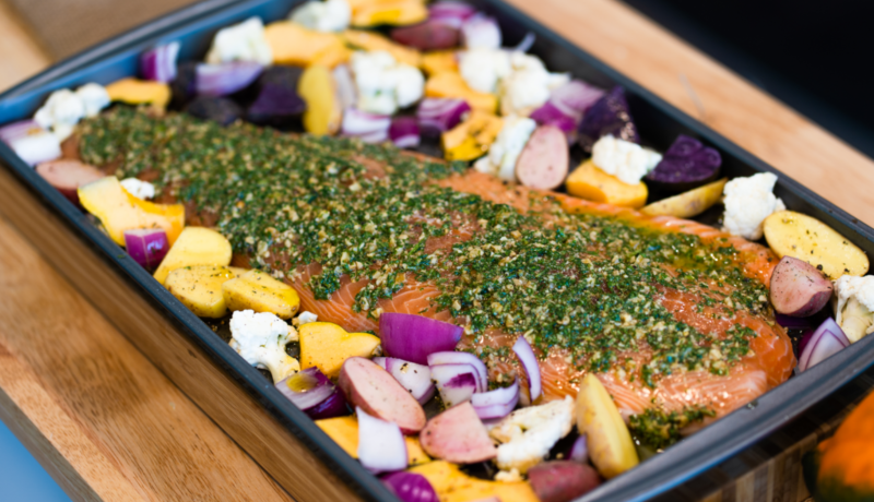A black baking sheet with chopped vegetables and a pesto-seasoned salmon filet. 