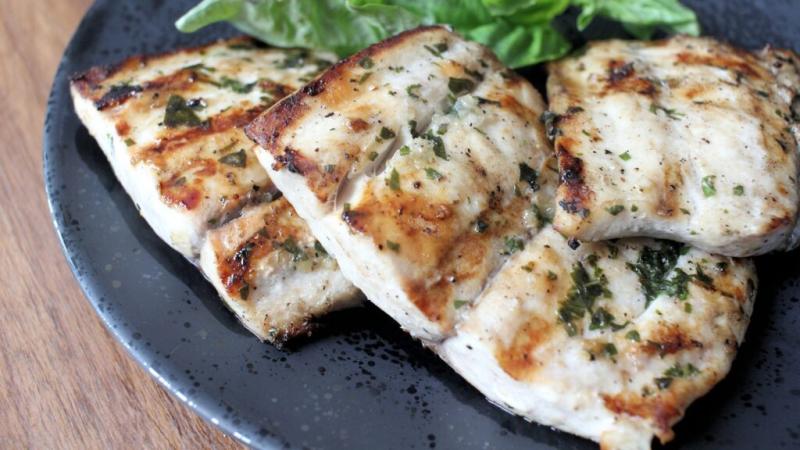 A black plate on a wooden table with three overlapping filets of seasoned white fish, grilled and garnished with lettuce.