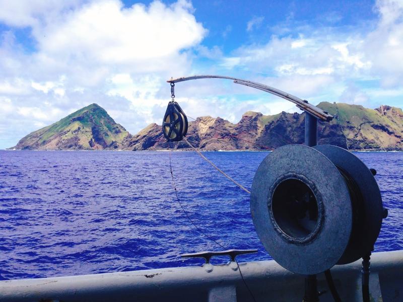 Close-up of a large reel and pulley on a ship, with blue water and jagged mountains in the background. 