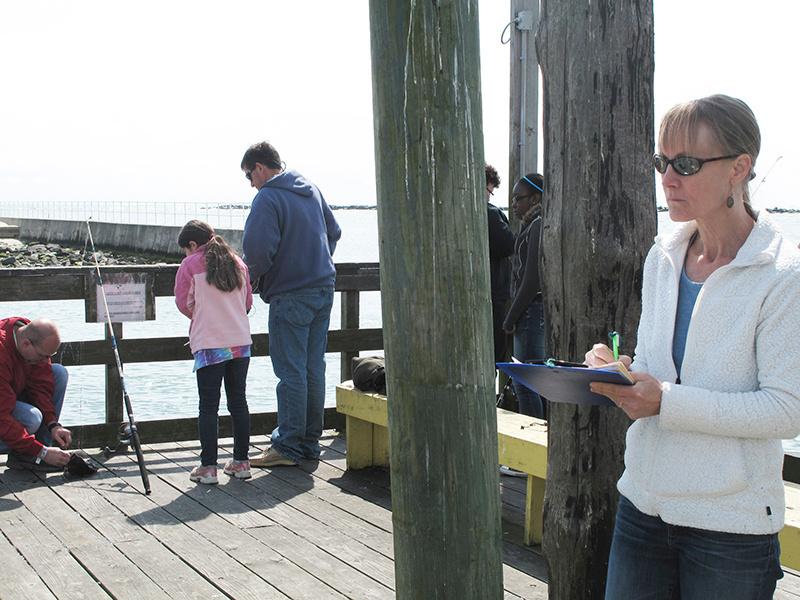Women in white jacket taking notes, family fishing in background. on a dock.