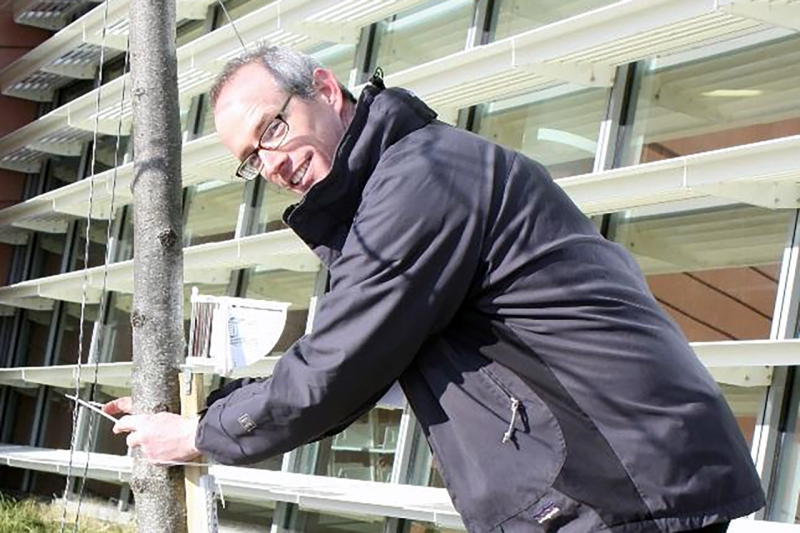 A man wearing a winter jacket looks at the camera as he leans over to secure a rain gauge to a tree
