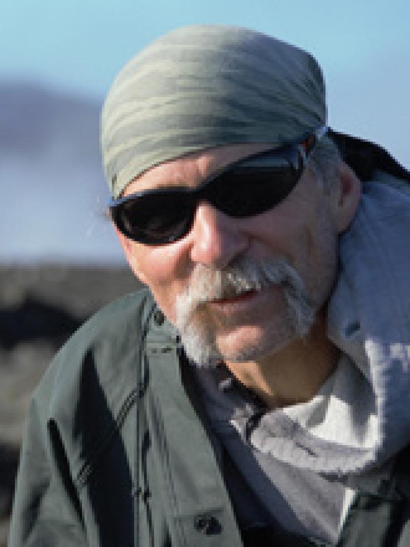 Man with bandana, glasses and mustache with a green coat, water and mountains in background. 