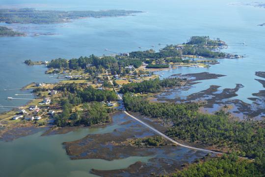 An aerial view of a small coastal community surrounded by water on all sides, accessed by a small road cutting through the water.