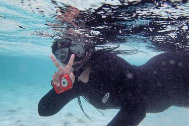 Gabi Clardy wears a mask, snorkel, and black dive skin while swimming at the surface of a shallow water area. There is sand beneath her and the water is crystal blue. She is looking at the camera and her right hand is giving the “peace” sign while holding a small red underwater camera. 