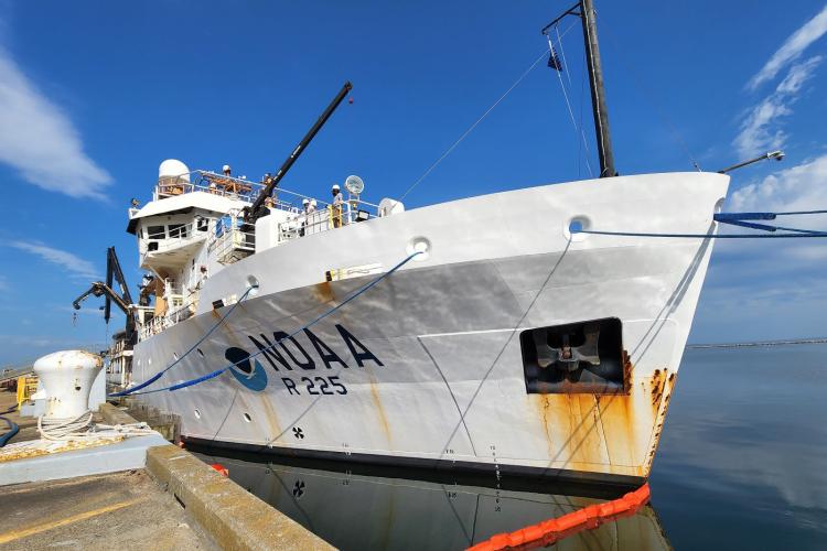 A color image taken on a sunny day. A large light colored ship with “NOAA” on the hull, The ship’s anchor is stowed midway up the bow, and rusty water has streaked from it down the hull. The ship’s bridge is visible along with cranes and pulleys mounted to the deck.  The water is calm and there are a few wispy clouds in the sky.