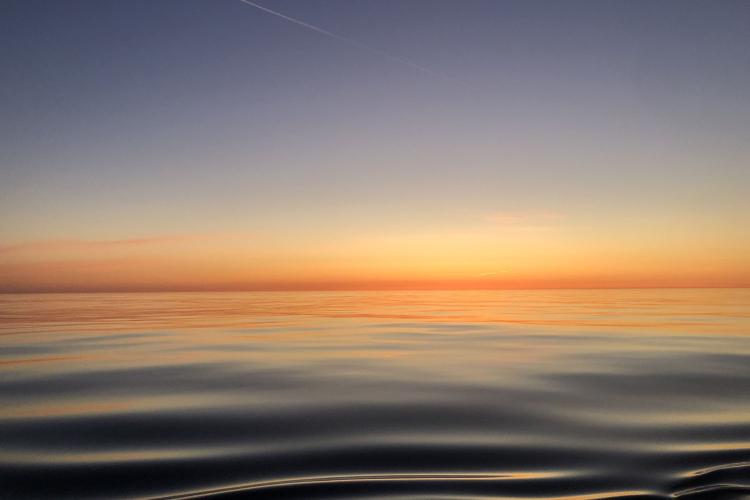 A color image taken at sunrise from the deck of a research ship. The water is calm and the rising sun’s light brightens the horizon at the image’s center.