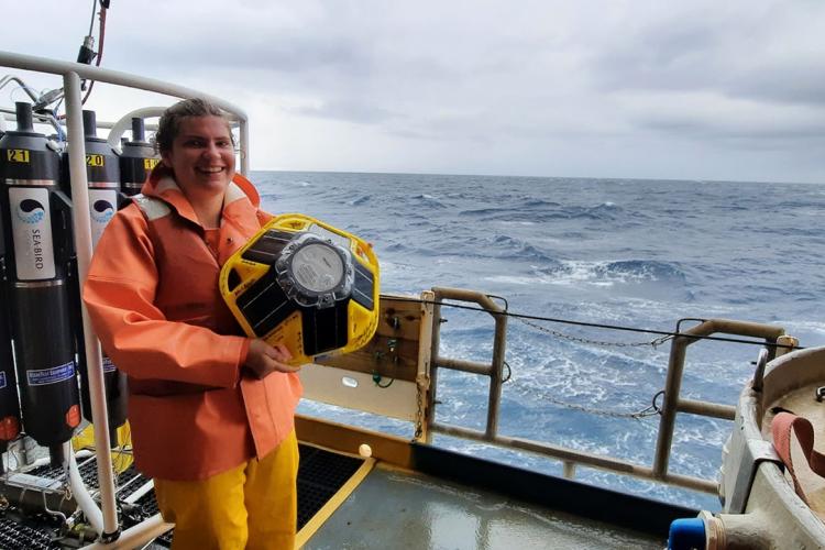 A woman in foul weather gear and an orange flotation vest stands on the ship’s deck. She holds a brightly-colored drifter buoy that is round, plump, and about the size of a beach ball.