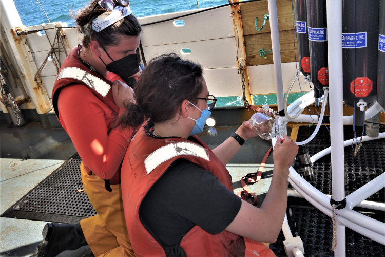 Color image taken on the deck of a research vessel during daylight. Two young women in foul weather gear and floatation vests crouch by a steel frame containing a series of bottles holding seawater samples. One woman holds a clear glass bottle that is connected by a hose to one of the bottles holding the sample. She is removing the water for further analysis.