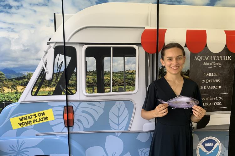 An undergraduate student stands in front of an photo booth exhibit with a kampachi fish prop. The photo booth background features a food truck on a Hawai'i roadside and showcases Hawai'i-grown aquaculture species on the pretend chalkboard menu, including mullet, oysters, and kampachi.
