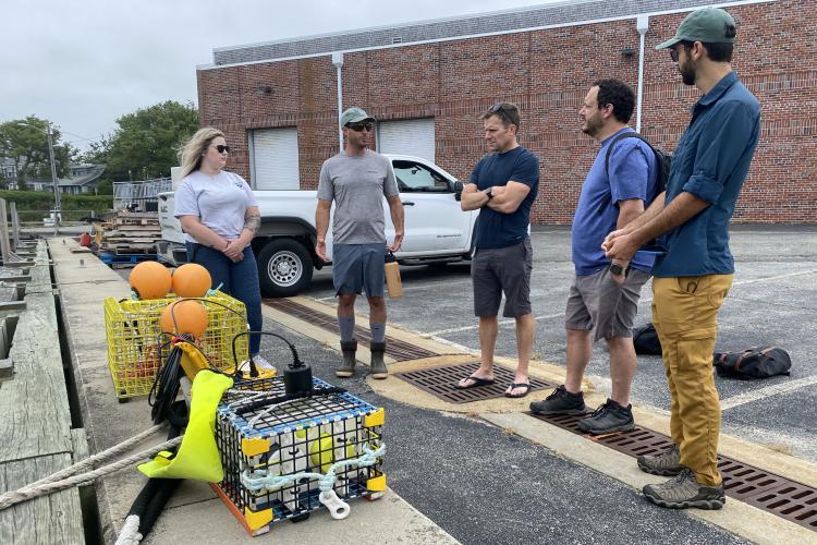 A color image taken in the summer during the day in overcast conditions. Two wire cages containing on-demand fishing gear are at left at the edge of the docking area that abuts the cement parking lot and lay-down area. The group is at the center. A brick building with two garage bays is in the background.