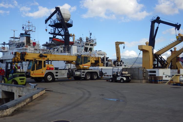 A photo taken pier-side with the NOAA Ship in the background, with a telehandler and forklift in the foreground.