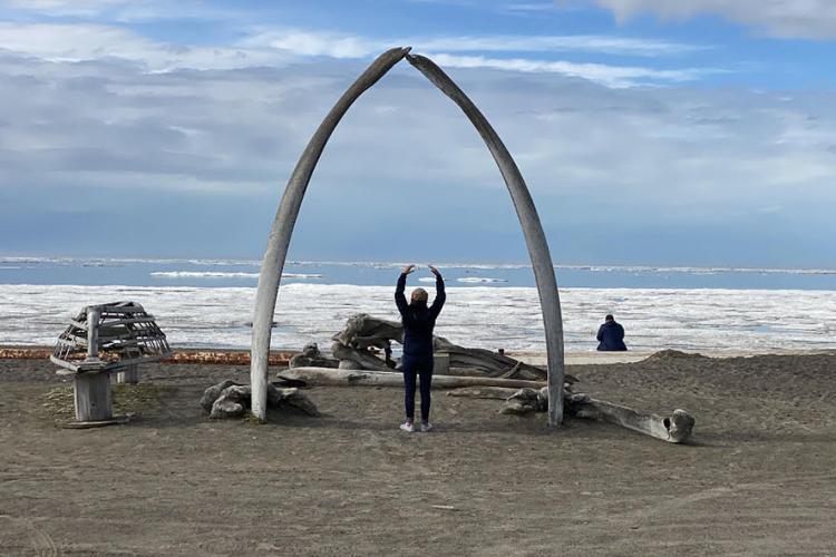 Mabel Baldwin-Schaeffer stands under an arch made of two whale ribs.  She has her arms raised, with her back to the camera and facing the Arctic Ocean on a beach in Utqiagvik, Alaska. The skeleton of a small overturned boat is propped up on the beach beside the arch. Sea ice gathers along the shore and in the distant offshore with a stretch of open water in between. Blue sky and gray clouds are visible above the horizon.