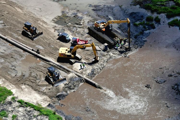 A pipeline delivers sediment to a marsh creation area. Construction equipment sits on the shoreline, and several construction workers wearing bright yellow safety vests monitor the progress.