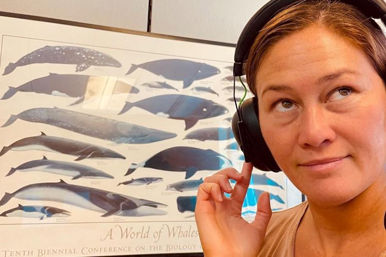 Cory Hom-Weaver looking up and to the side while holding her finger to her headphones over her head. In the background there is a framed poster of whales of the world