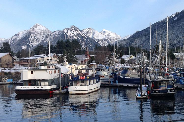 Photo showing fishing boats docked in the foreground, buildings in the middle ground, and snowy mountains in the background under blue skies.