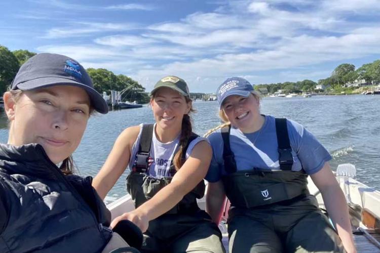 Three female researchers sit on a boat with the water and blue sky behind them. 