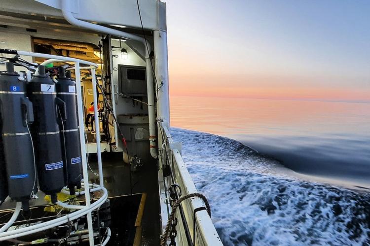 On the left, the deck of the NOAA Ship Henry B. Bigelow with electrical wiring and part of a conductivity temperature and depth rosette water sampler visible, including three gray cylindrical bottles for sampling seawater. On the right, the calm flat and blue ocean over the deck of the ship with an orangish pink sunset on the horizon.